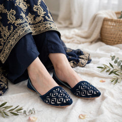 A person wearing navy blue flat shoes with white geometric patterns, seated on a white textured blanket with a navy and gold embroidered garment draped over the lap, and a woven basket in the background.
