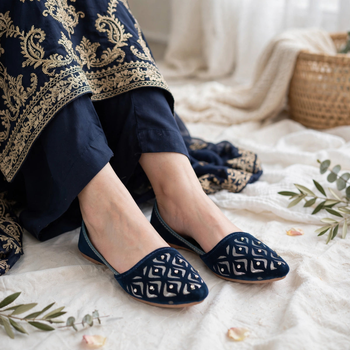 A person wearing navy blue flat shoes with white geometric patterns, seated on a white textured blanket with a navy and gold embroidered garment draped over the lap, and a woven basket in the background.